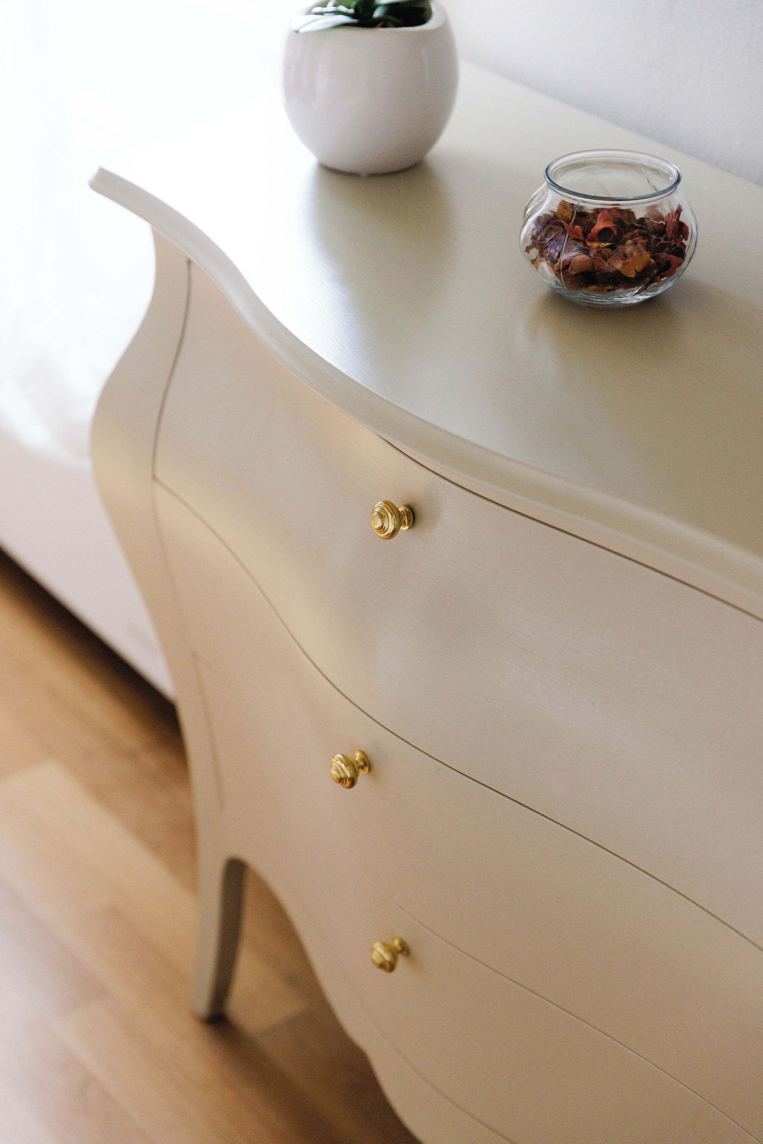 A white dresser with gold knobs and a glass bowl containing dried flowers on top.