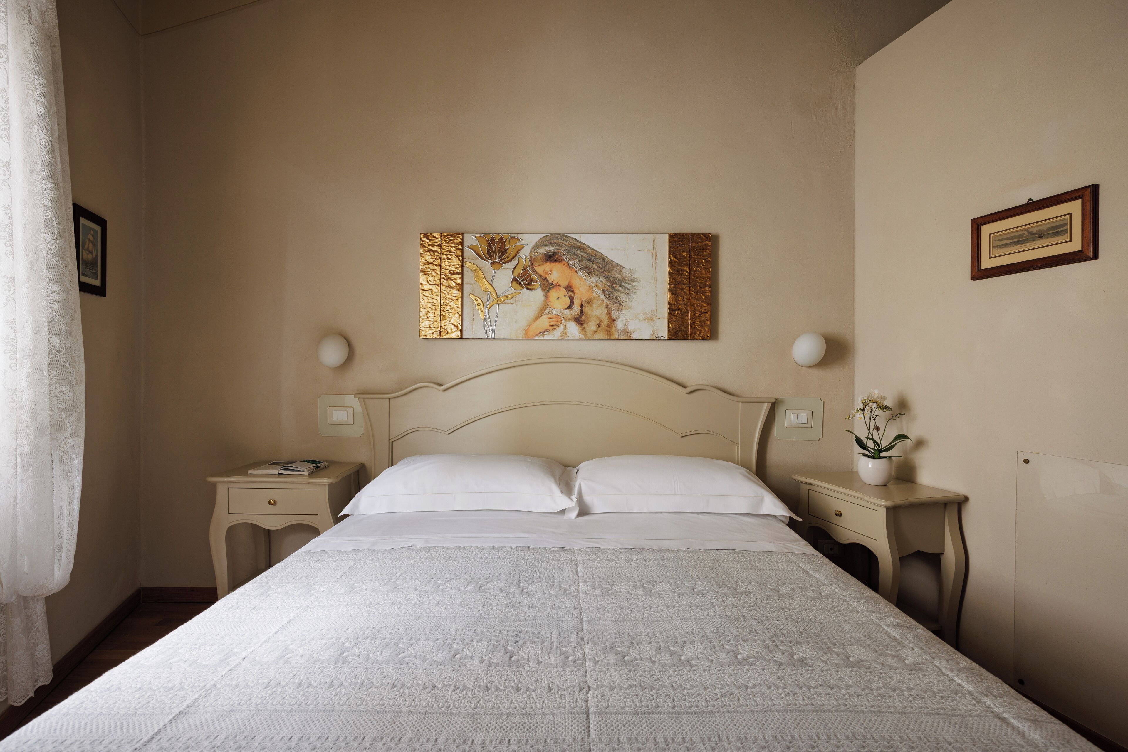 A neatly made bed with a white bedspread, two bedside tables with lamps, a framed picture on the wall, and a small potted plant on one of the nightstands.