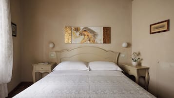 A neatly made bed with a white bedspread, two bedside tables with lamps, a framed picture on the wall, and a small potted plant on one of the nightstands.