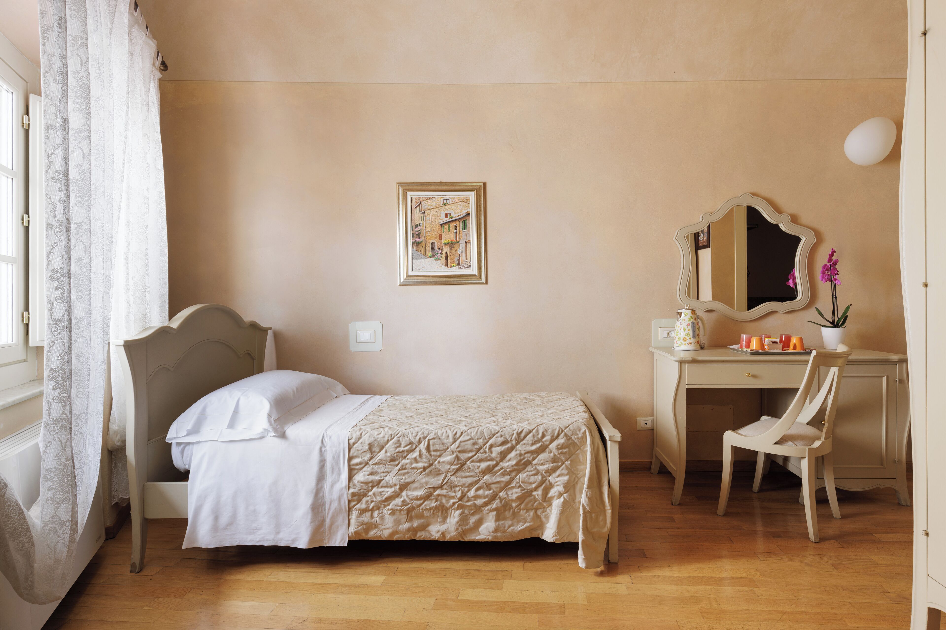 Un dormitorio con cama, escritorio, silla, espejo, un cuadro en la pared y ventana con cortinas.