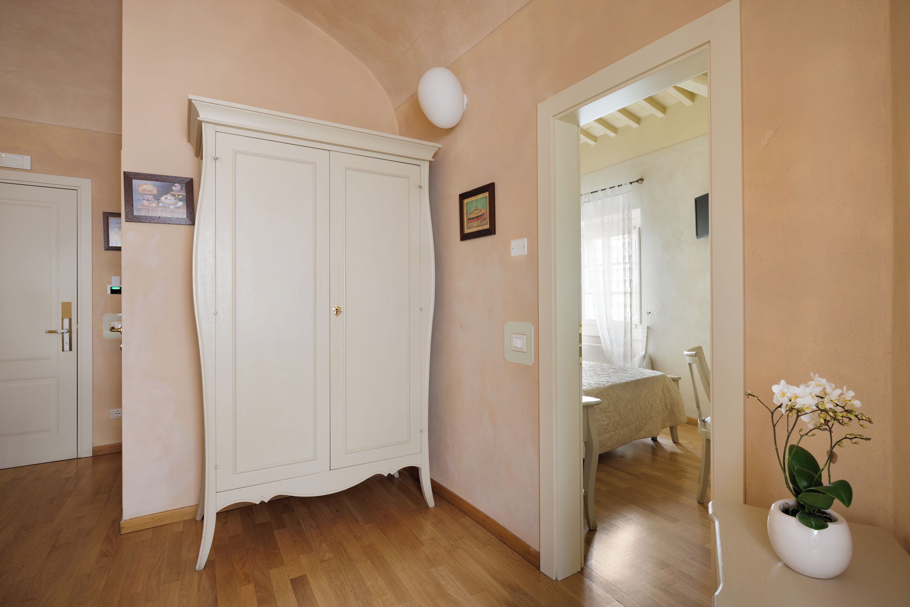 Una habitación con un armario blanco, un jarrón blanco con flores y una cama al fondo.