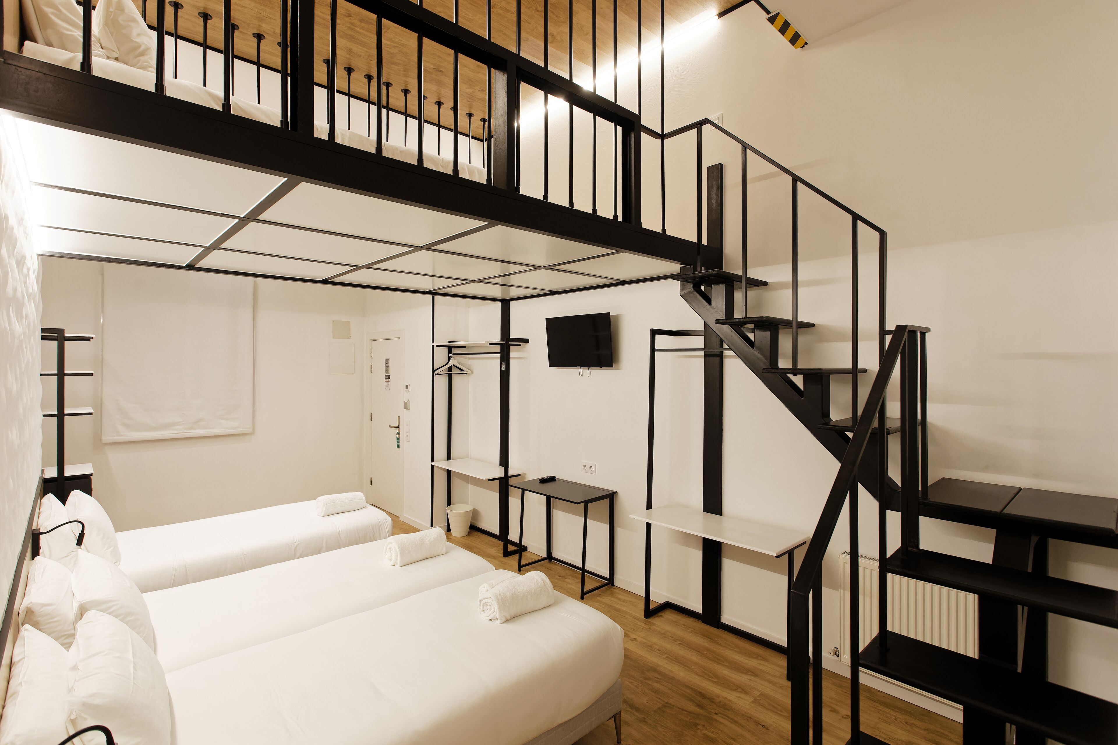 Hotellihuone, jossa on kaksi sänkyä, puulattia, musta portaikko ja seinälle asennettu televisio.