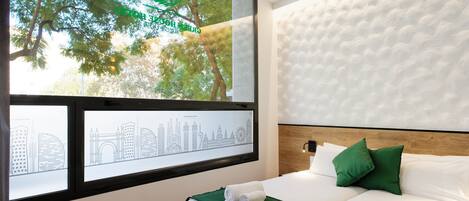 A hotel room with a bed, green pillows, a white headboard, and a window with a cityscape view.