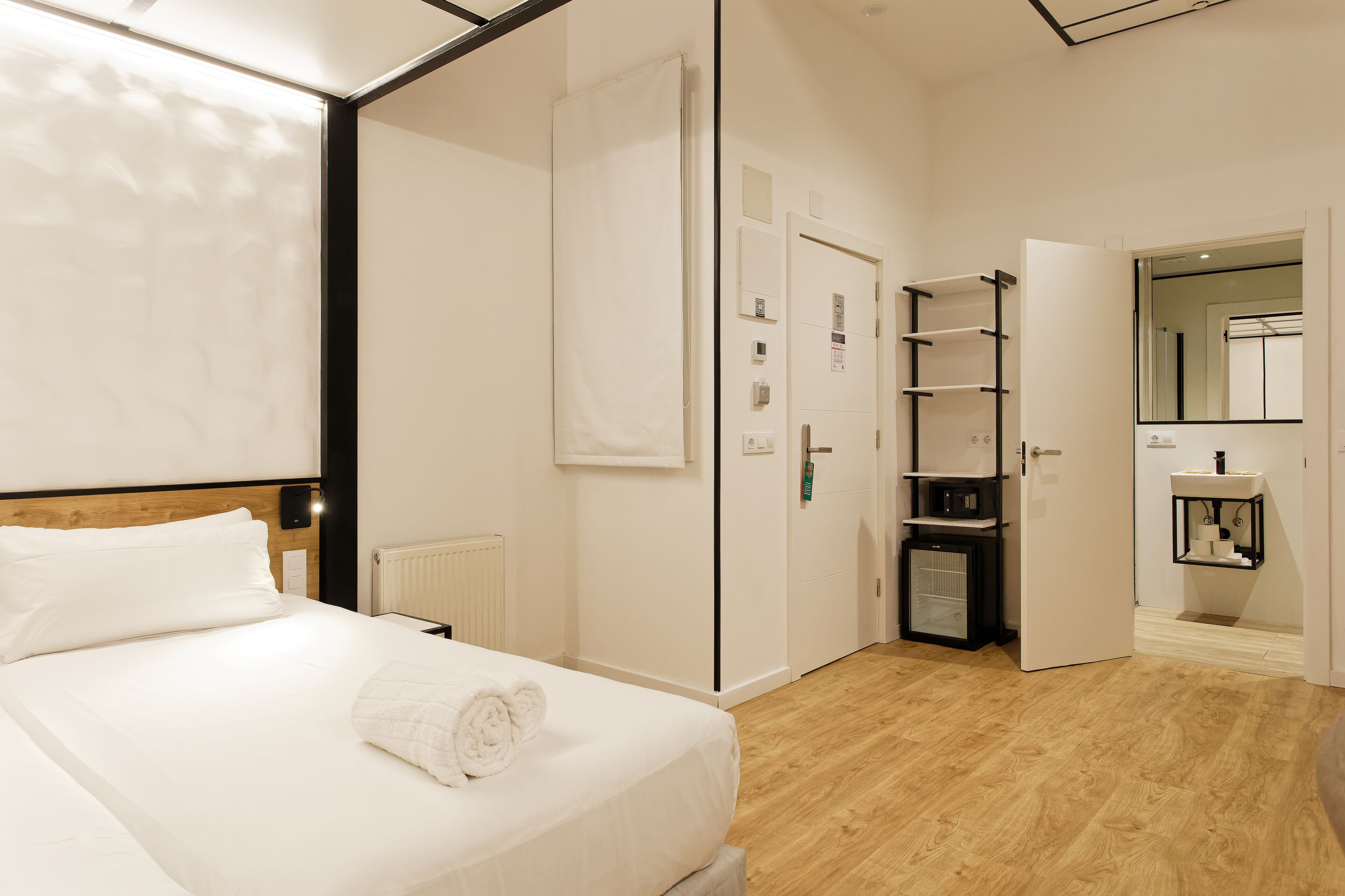 A hotel room with a bed, a radiator, a wardrobe, a shelf unit, and a bathroom mirror.