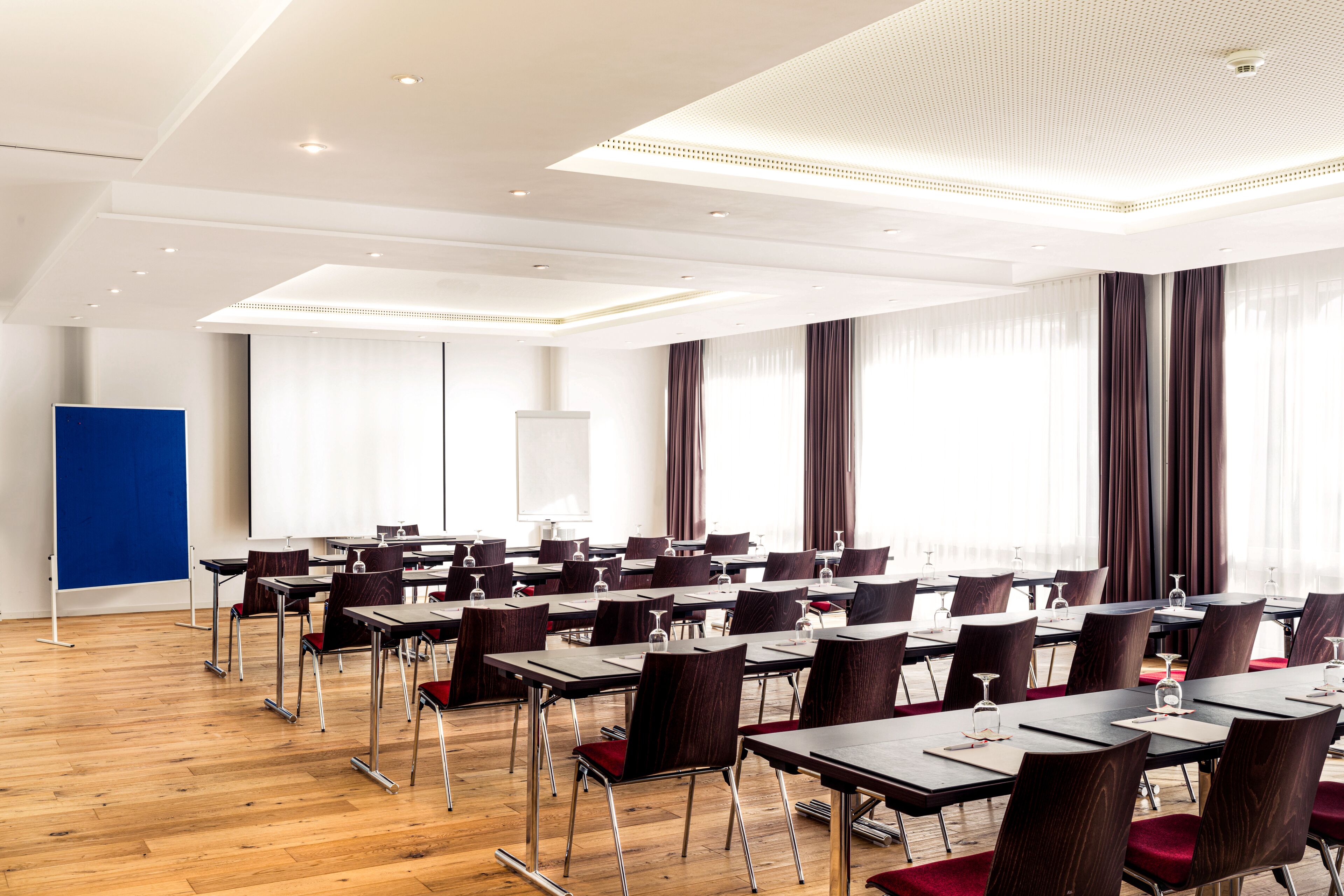 Ein Konferenzraum mit mehreren Tischen und Stühlen, Holzboden und großen Fenstern mit Vorhängen.
