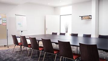 A conference room with a long table, six chairs, a whiteboard, and a window with a view of bare branches.
