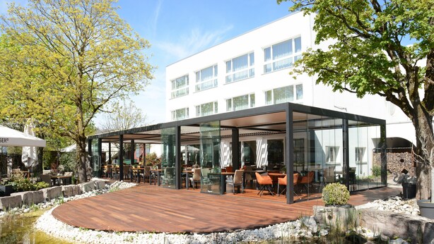 Un espacio moderno para comer al aire libre con terraza de madera, cerramiento de cristal y un estanque.