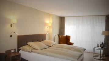 A hotel room with a large bed, two bedside lamps, a chair, a small table with a plant, and a window with curtains.