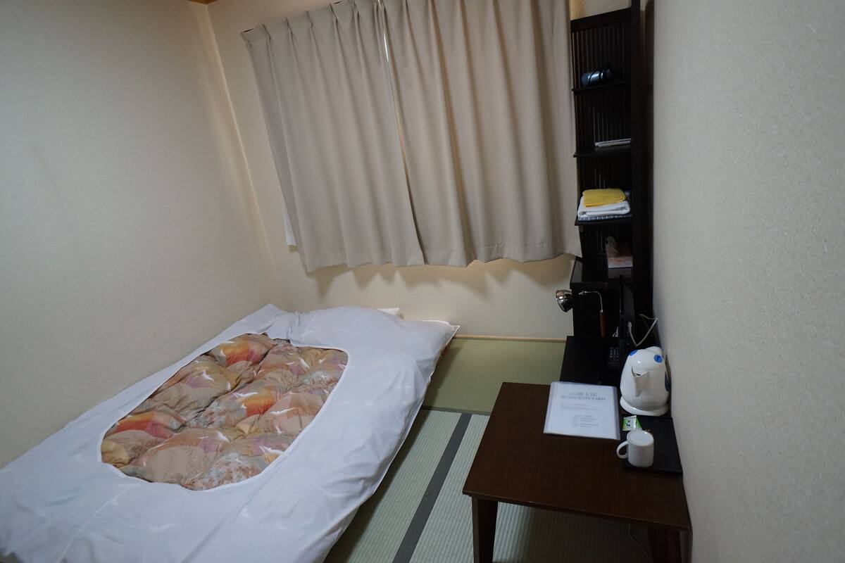 A small hotel room with a single bed, a bedside table with a coffee maker, a shelf with towels, and a window with curtains.