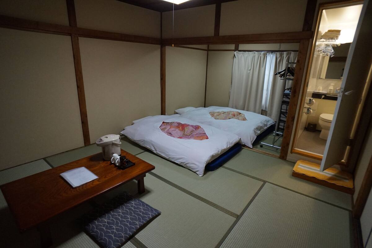 A traditional Japanese-style room with tatami flooring, a wooden table with a cushion, two beds with pillows, a small table with a telephone, and a bathroom visible through a sliding door.