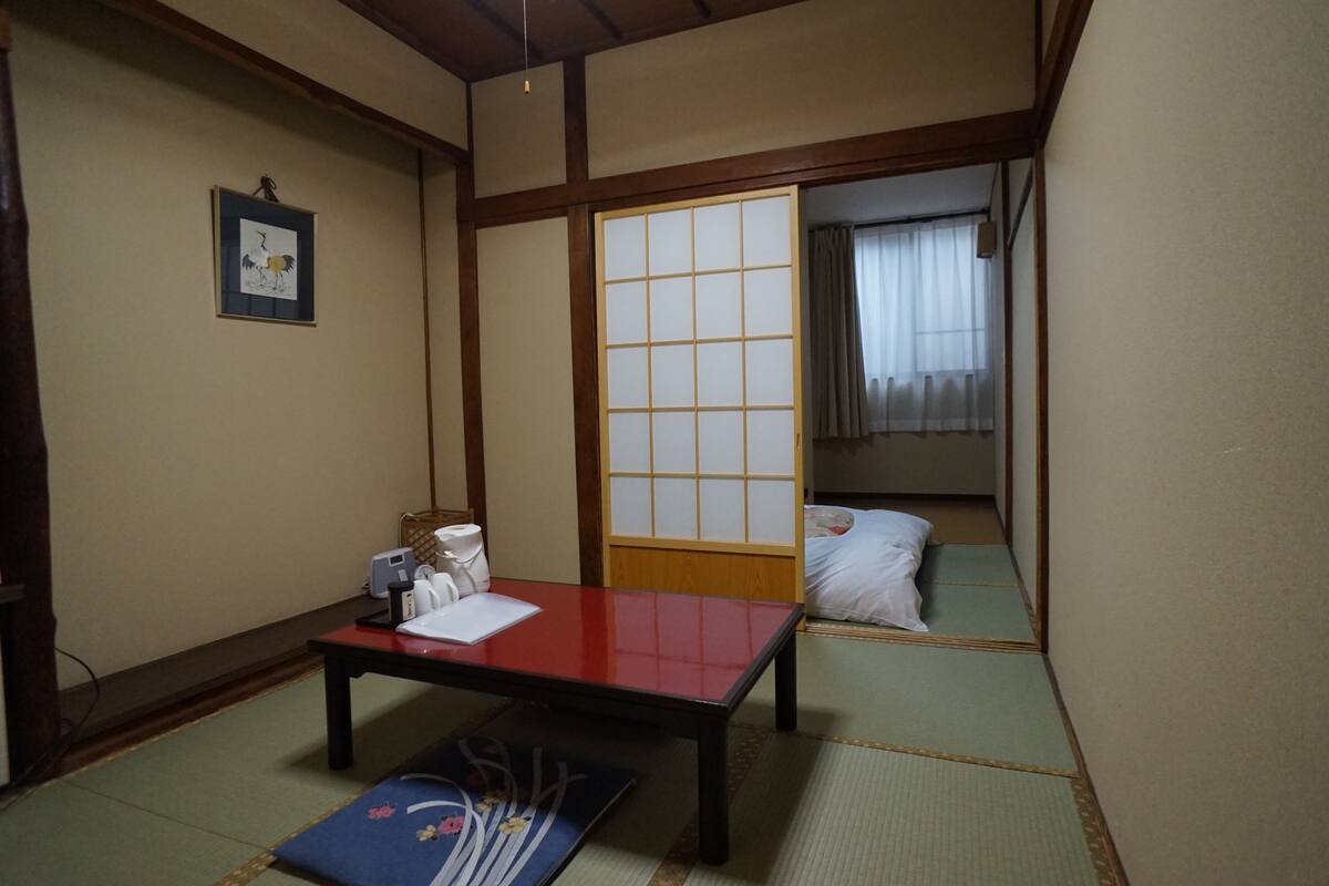A traditional Japanese room with tatami flooring, a low wooden table, a shoji screen door, and a bed with white bedding.