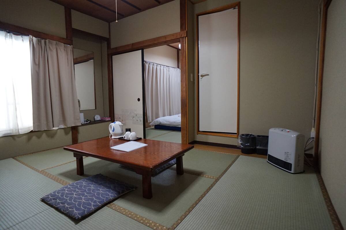 A traditional Japanese room with tatami flooring, a wooden table with a cushion, a heater, and a sliding door leading to a bed.