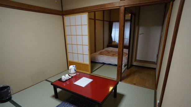 A traditional Japanese room with tatami flooring, a low wooden table with a red top, a small bench, and a sliding door leading to a bedroom with a bed and a nightstand. - Ryokan Katsutaro (Tokyo)
