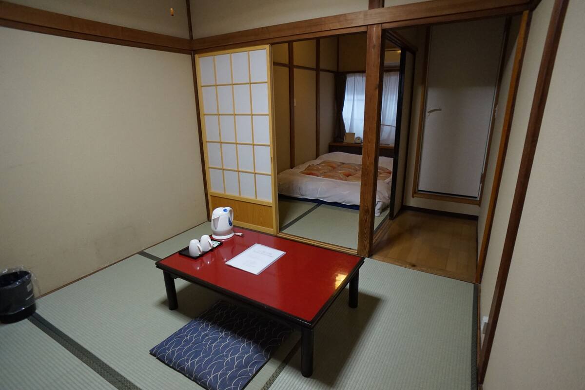 A traditional Japanese room with tatami flooring, a low wooden table with a red top, a small bench, and a sliding door leading to a bedroom with a bed and a nightstand.