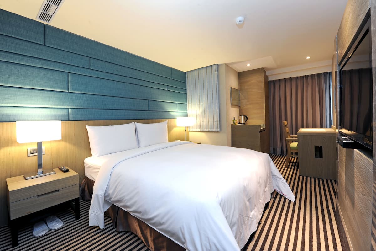 A hotel room with a large bed, bedside tables with lamps, a small desk, a chair, a minibar, and a view of the city.
