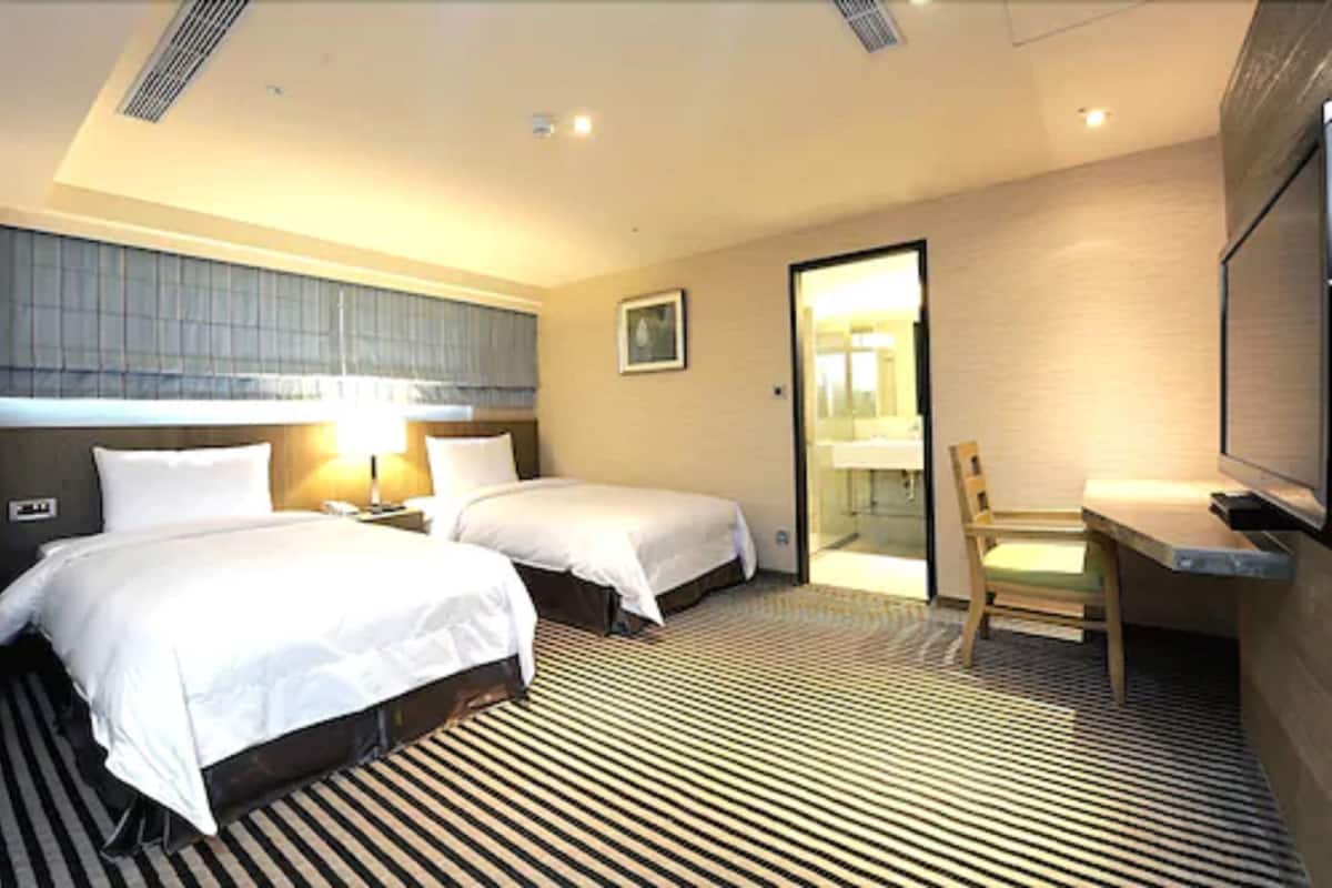 A hotel room with two beds, a desk, a chair, a television, and a bathroom visible through an open door.