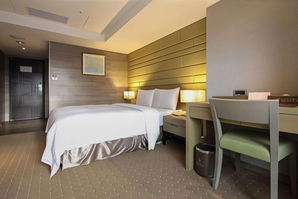 Ein Hotelzimmer mit Bett, Nachttisch, Lampe, Stuhl und Mülleimer.