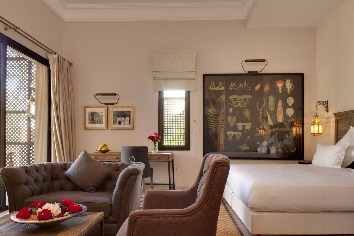 Quarto de hotel com cama, duas poltronas, escrivaninha com cadeira, mesa de jantar e um arranjo de flores, além de uma grande obra de arte na parede.
