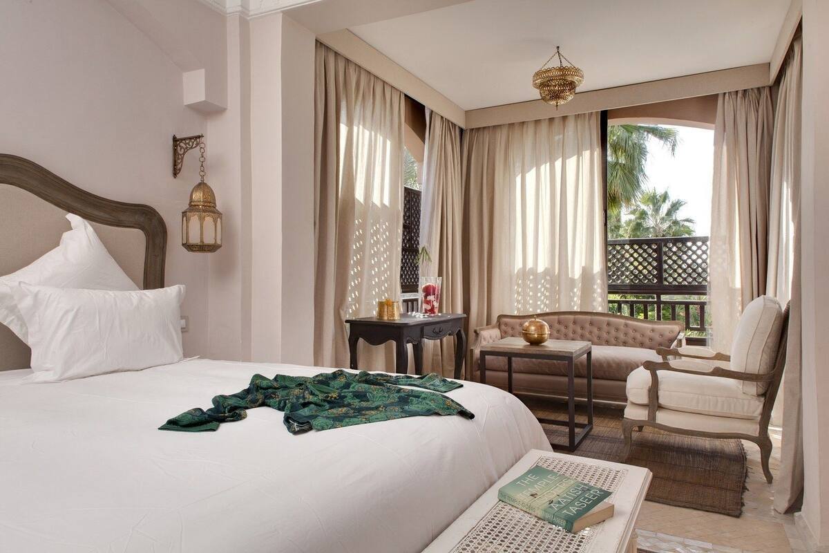 Quarto de hotel com uma cama grande, uma mesa pequena, uma cadeira e varanda com cortinas.