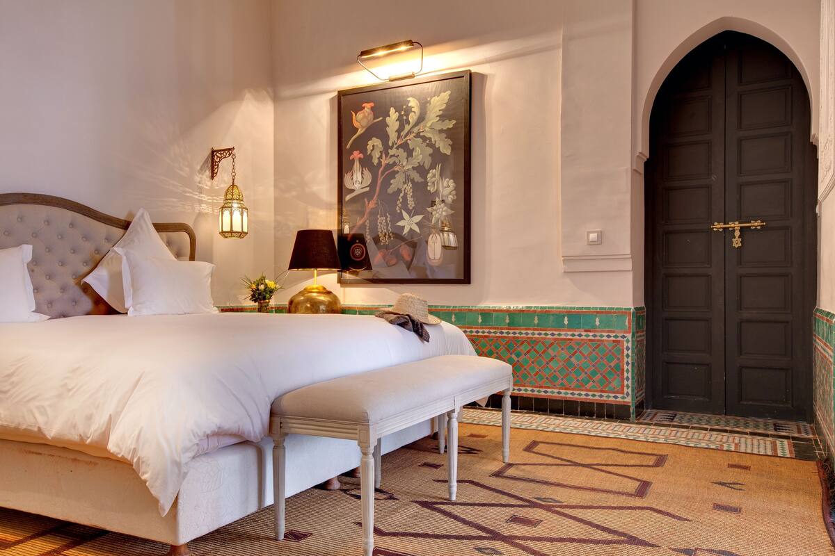 Quarto com cama, banco, luminária na parede e uma porta.