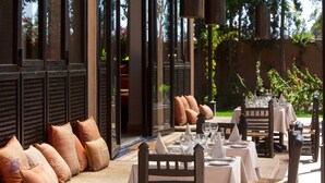 Un área de comedor al aire libre con mesas dispuestas para dos, asientos acolchados y vistas al jardín.
