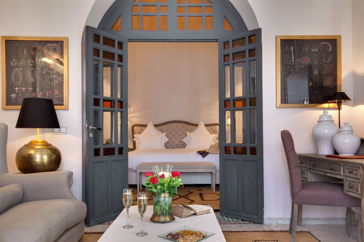 Quarto de hotel com cama, escrivaninha, cadeira, mesa com um vaso de flores e duas taças de vinho.