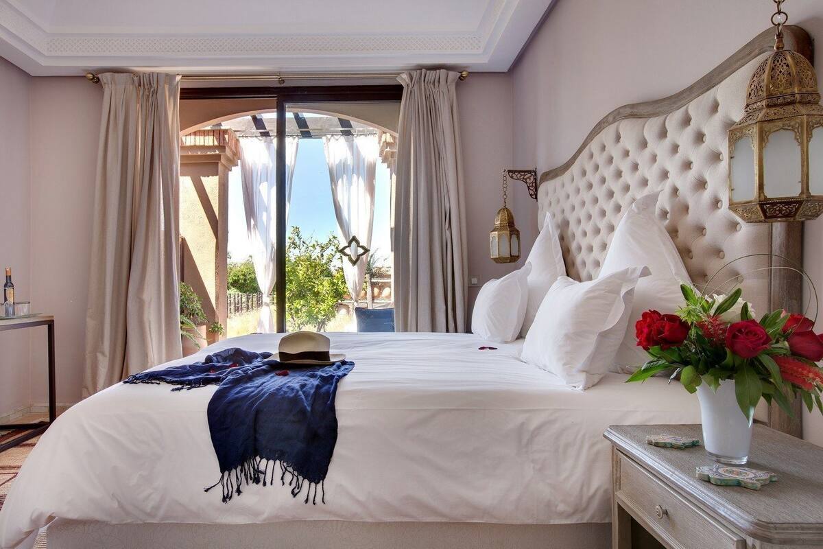 Quarto de hotel com uma cama grande, vista para o exterior, luminária decorativa e um vaso com flores.