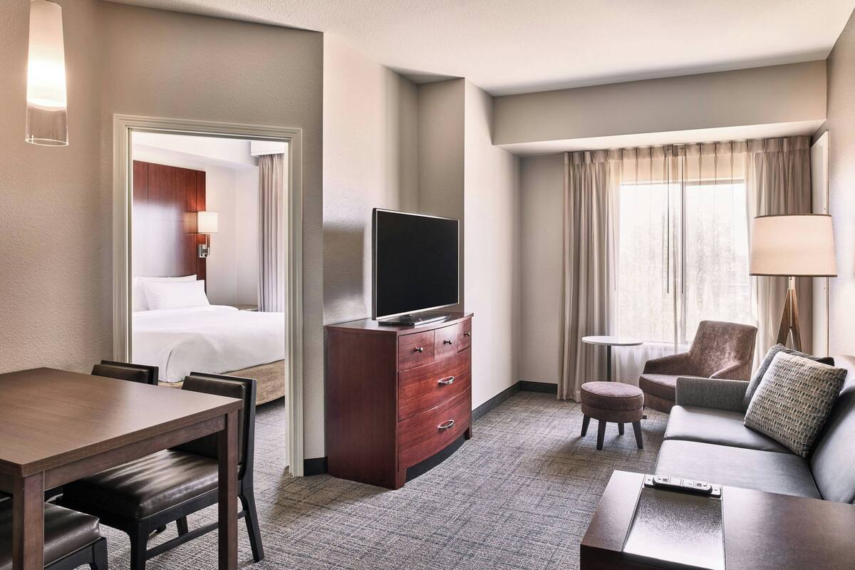 A hotel room with a bed, a TV on a wooden dresser, a dining table with chairs, a sofa, and a small round table.