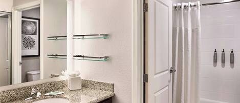 A modern bathroom with a granite countertop, a large mirror, a glass shelf, a white bathtub, and a shower curtain.