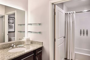 A modern bathroom with a granite countertop, a large mirror, a glass shelf, a white bathtub, and a shower curtain.