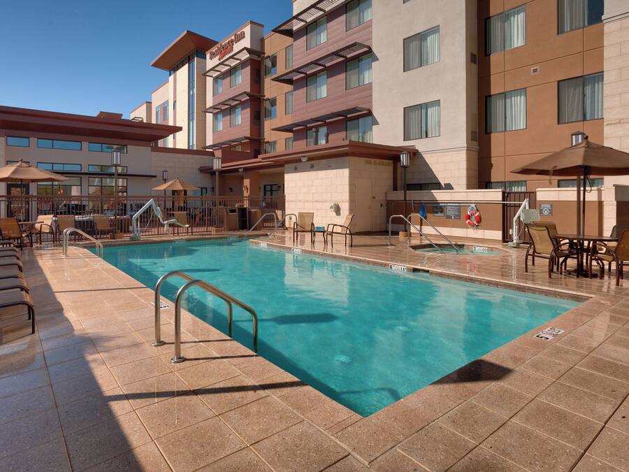 Zona de piscina de hotel con múltiples sillas de descanso y sombrillas, una piscina azul cristalina y un edificio de varios pisos y ventanas.