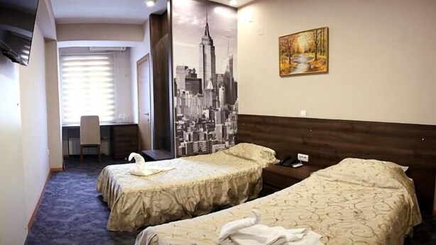 A hotel room with two beds, a cityscape mural, a painting, and a desk with a chair. - Ambasador (Skopje)