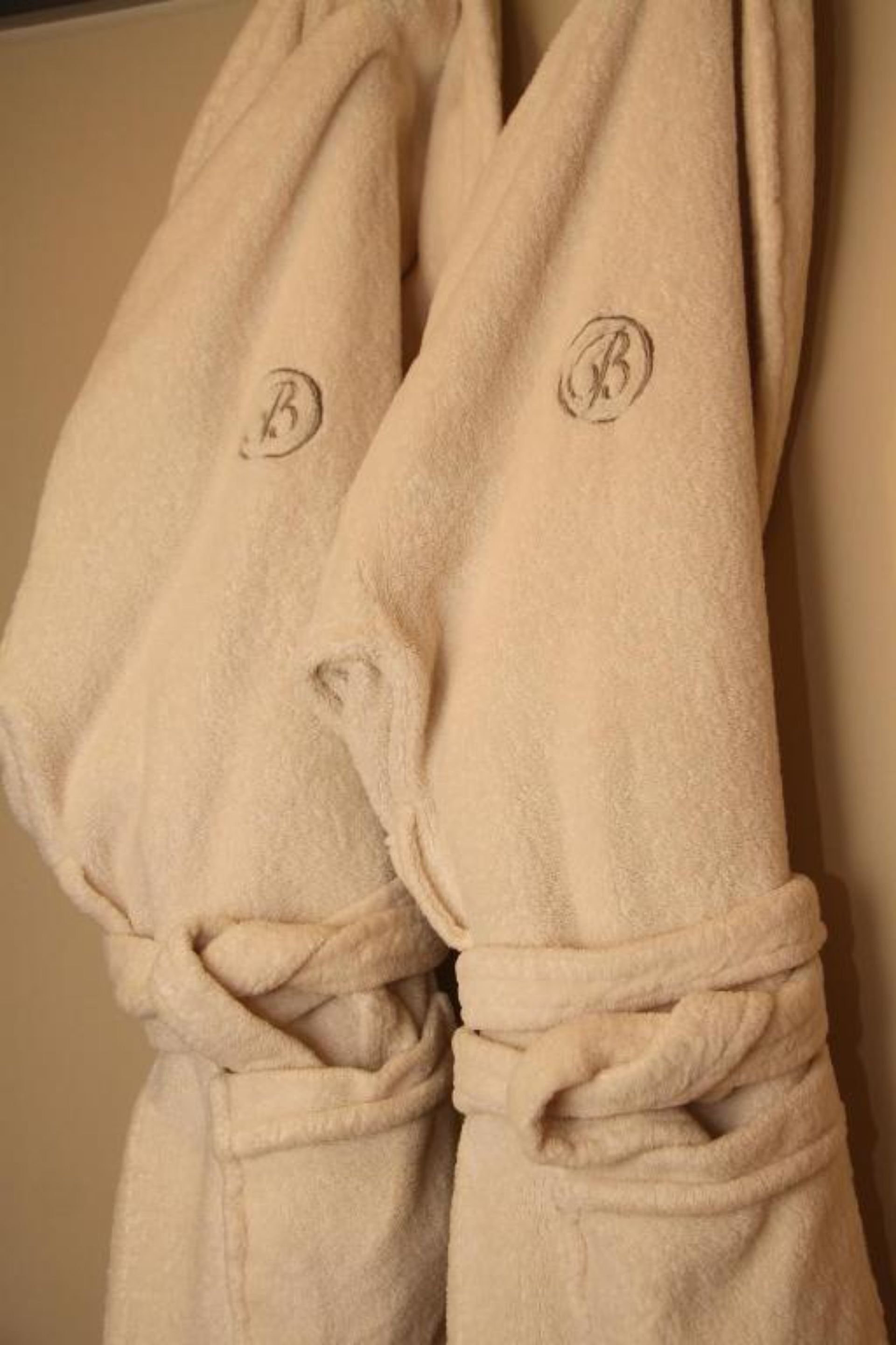 Two beige bathrobes with embroidered initials hanging on a wall.