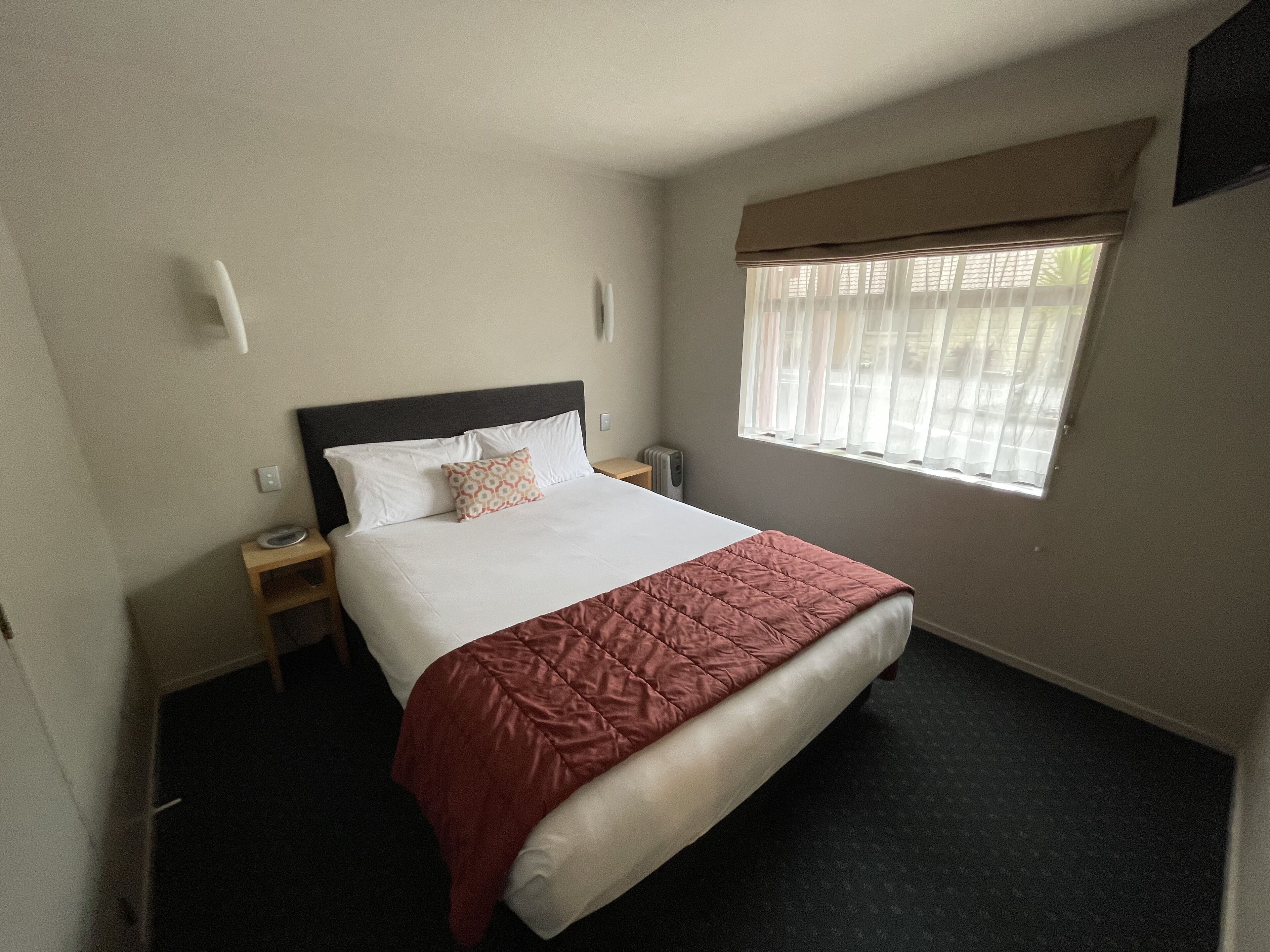A hotel room with a bed, a window with curtains, a bedside table, and a wall-mounted light.