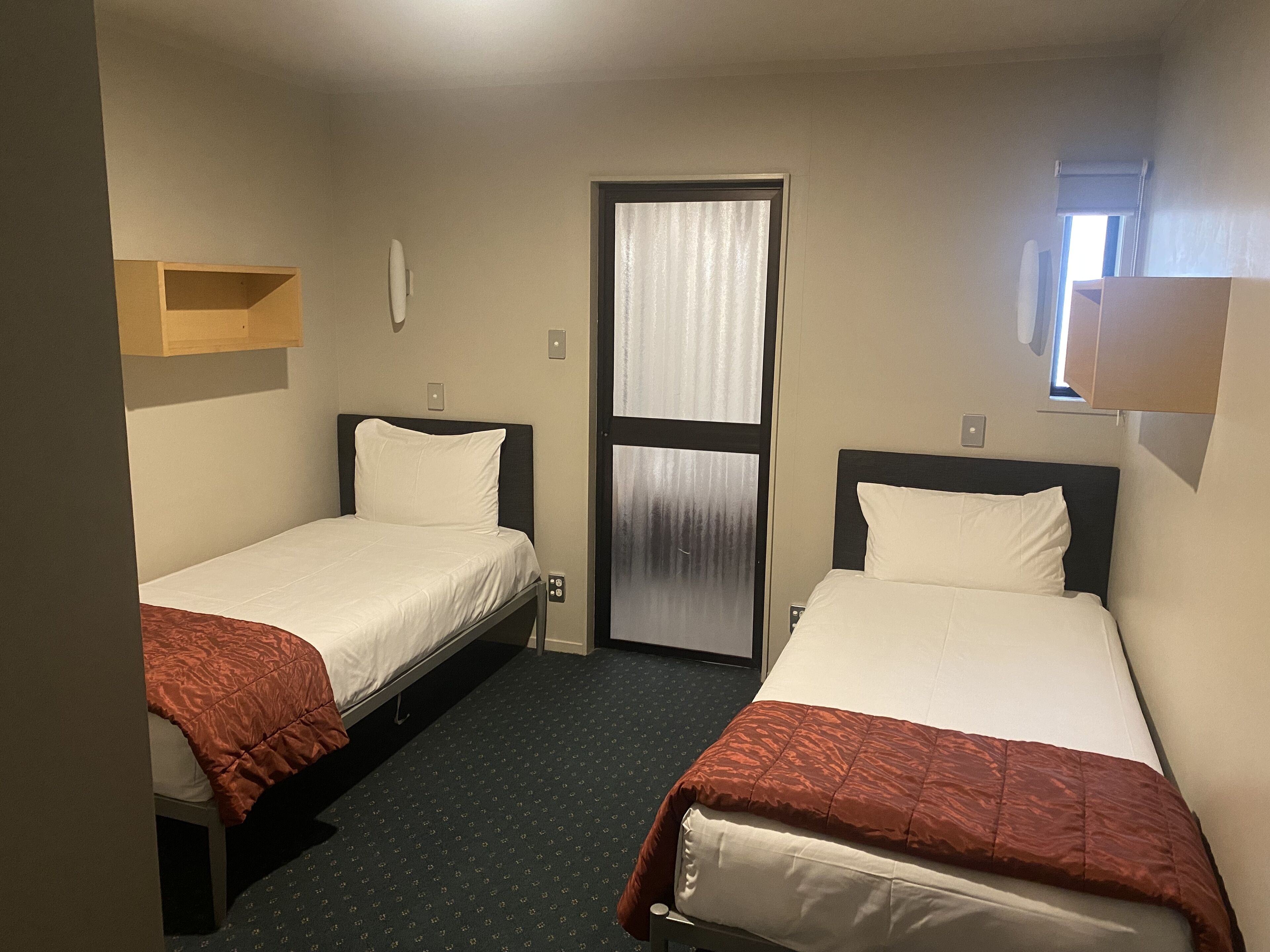 A hotel room with two single beds, a window with curtains, a door, and a small shelf above one bed.
