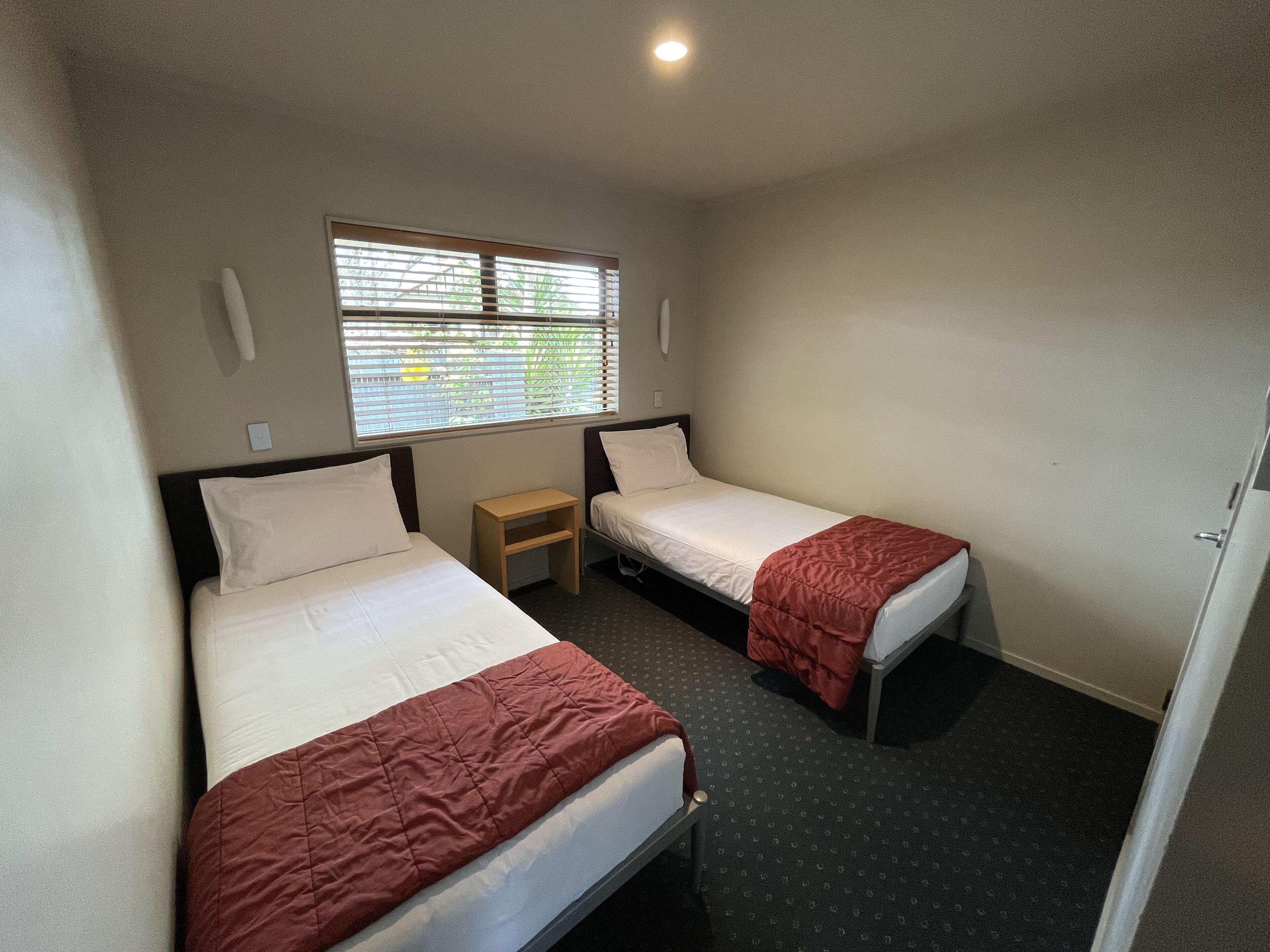A small, clean room with two single beds, a small table, and a window with blinds.