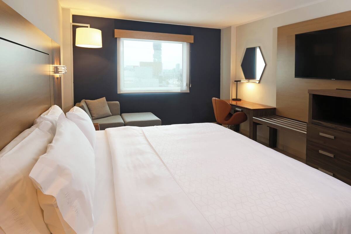 A hotel room with a large bed, a desk with a chair, a TV, and a window with a view of the city.