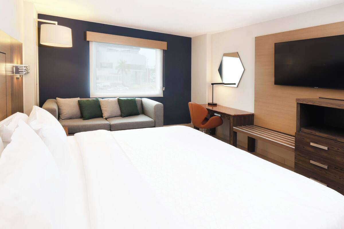 A hotel room with a large bed, a sofa, a desk, a chair, a TV, and a window with a view of buildings.