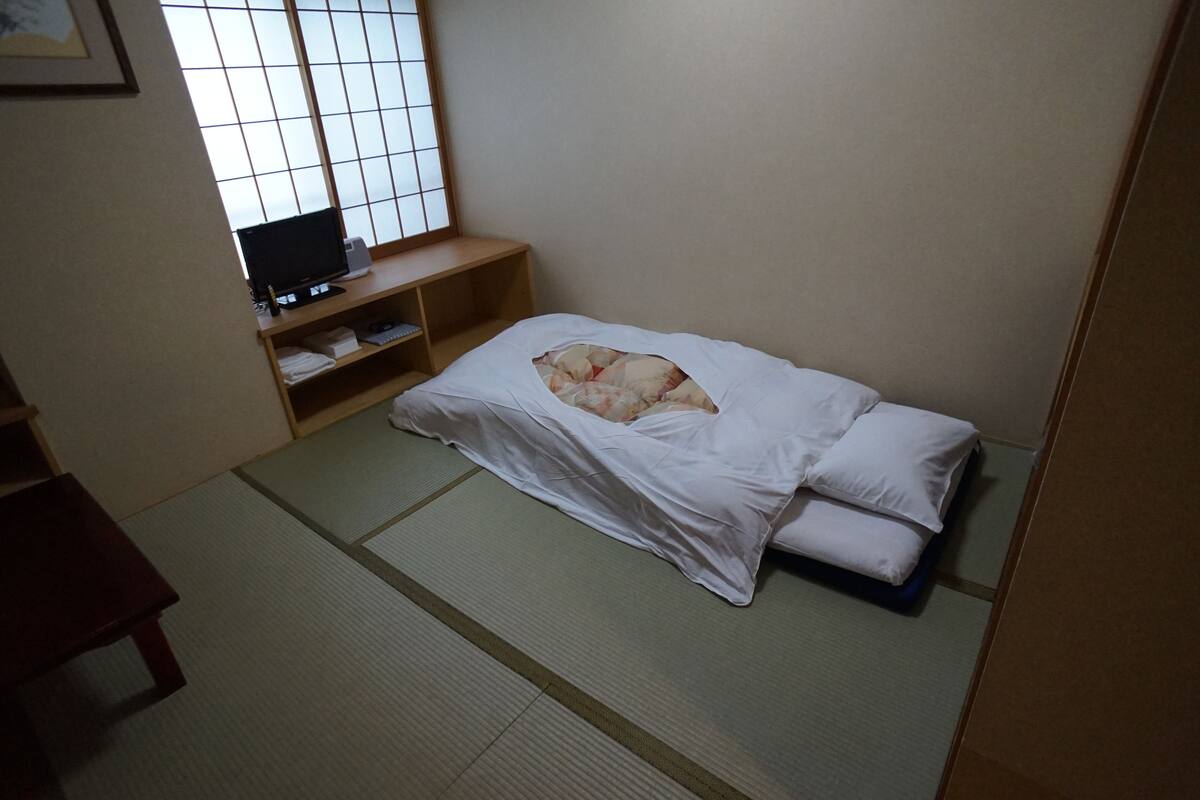 A room with tatami flooring, a bed with white bedding, a TV on a wooden shelf, and a window with a grid pattern.