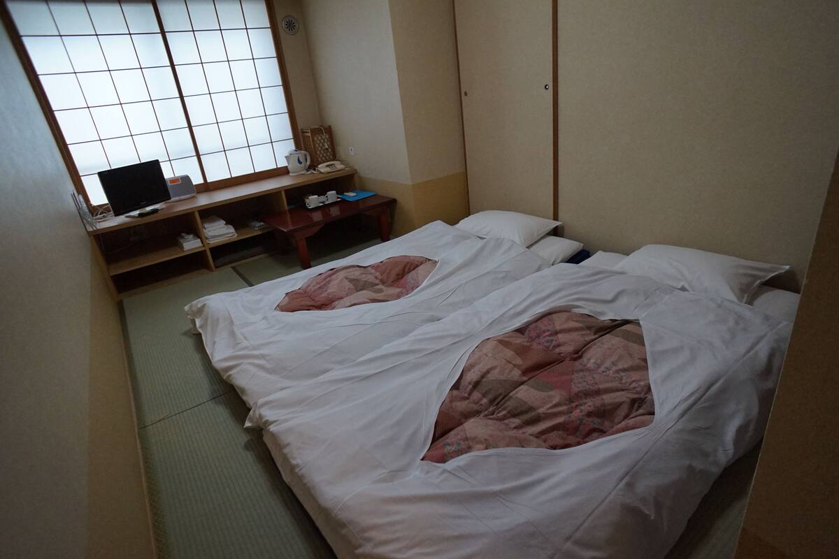 A traditional Japanese room with tatami flooring, two beds with white bedding, a desk with a laptop, and a shoji screen.