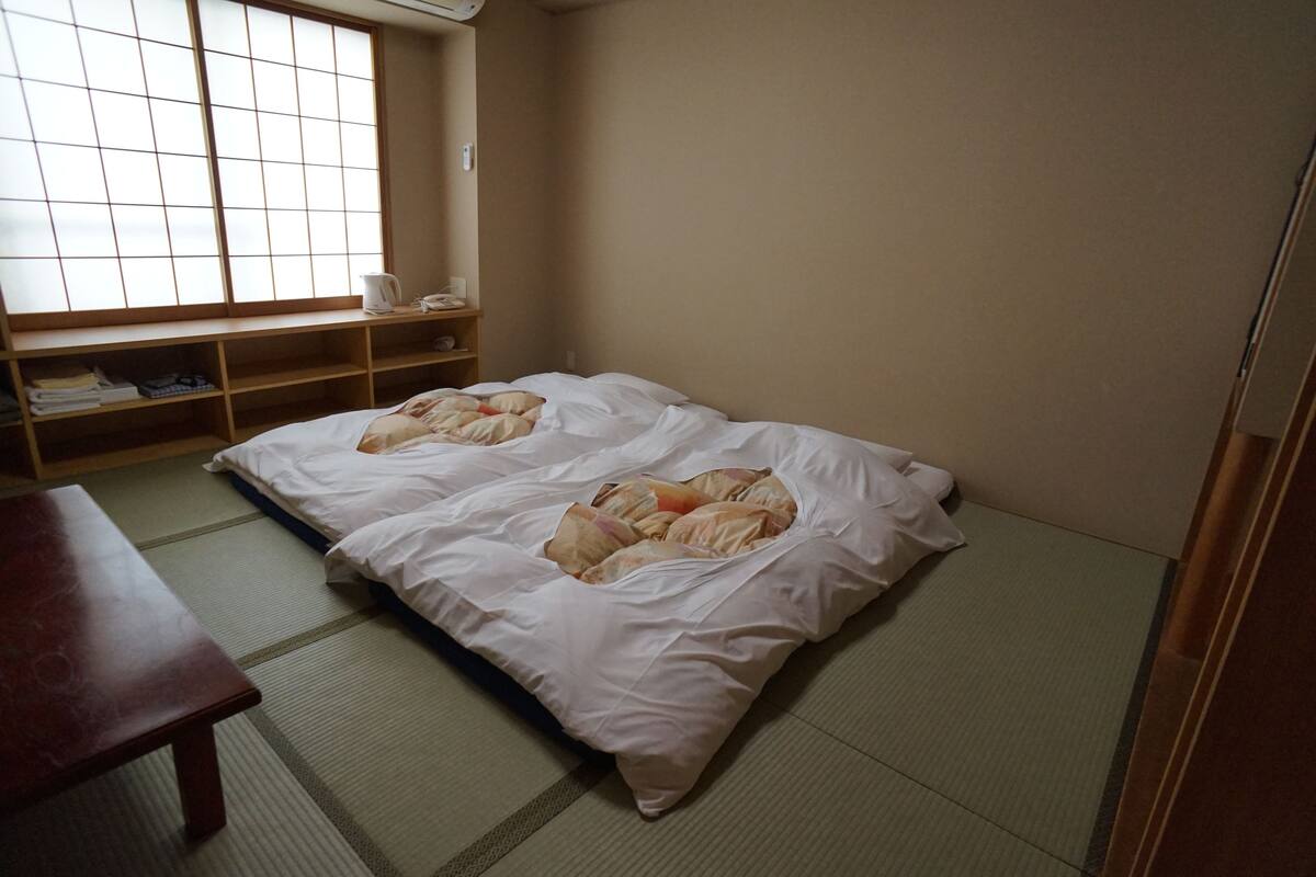 A room with tatami flooring, a futon bed, a wooden table, and a shoji screen window.