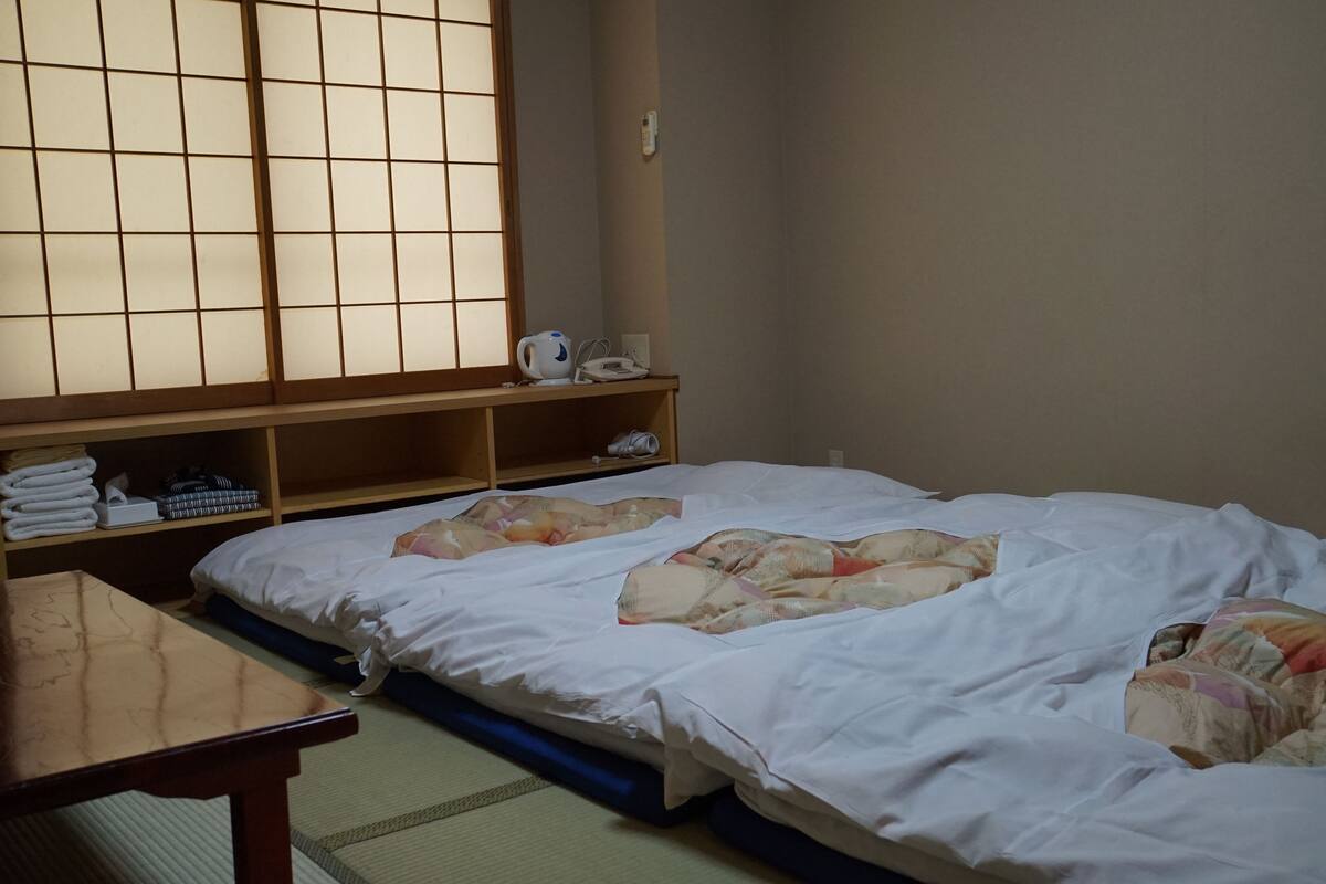 A room with a bed, a wooden table, and a shelf with towels and a kettle.