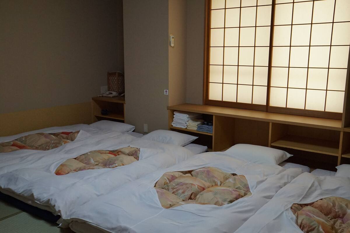 A hotel room with three beds, a wooden shelf with folded towels, and a large window with a grid pattern.