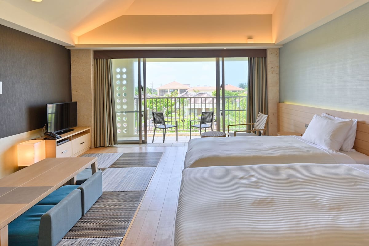 A hotel room with a large bed, a TV on a wooden console, a balcony with chairs, and a view of buildings.