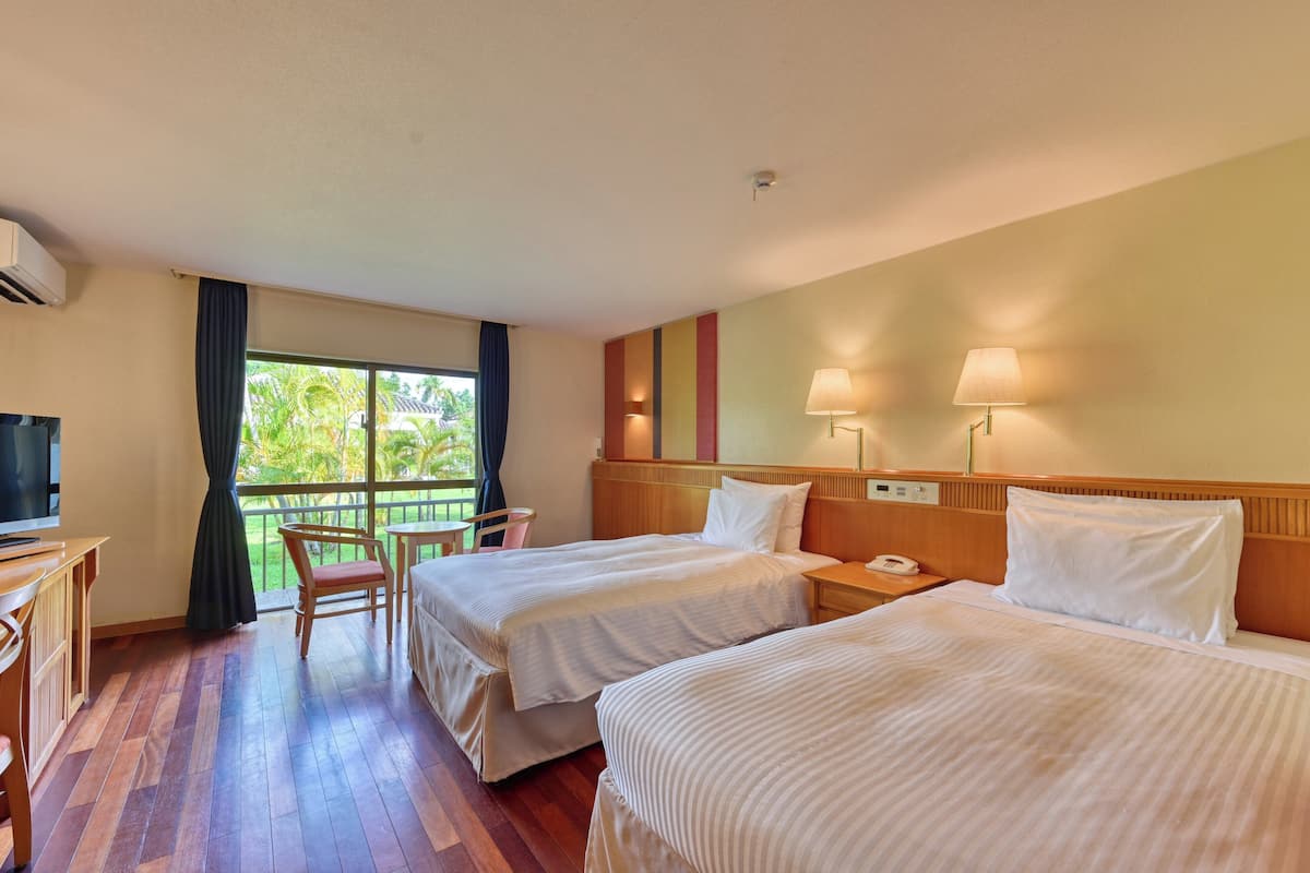 A hotel room with two beds, a desk, a chair, a TV, and a balcony with a view of greenery.