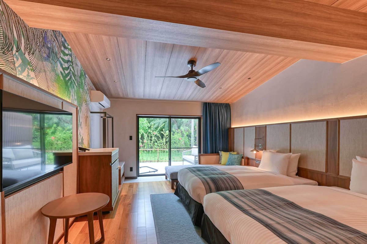 A modern bedroom with two beds, a ceiling fan, and a large window overlooking greenery.