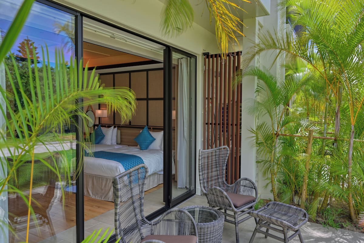 A modern bedroom with a glass door leading to a balcony, wicker furniture, and lush greenery.