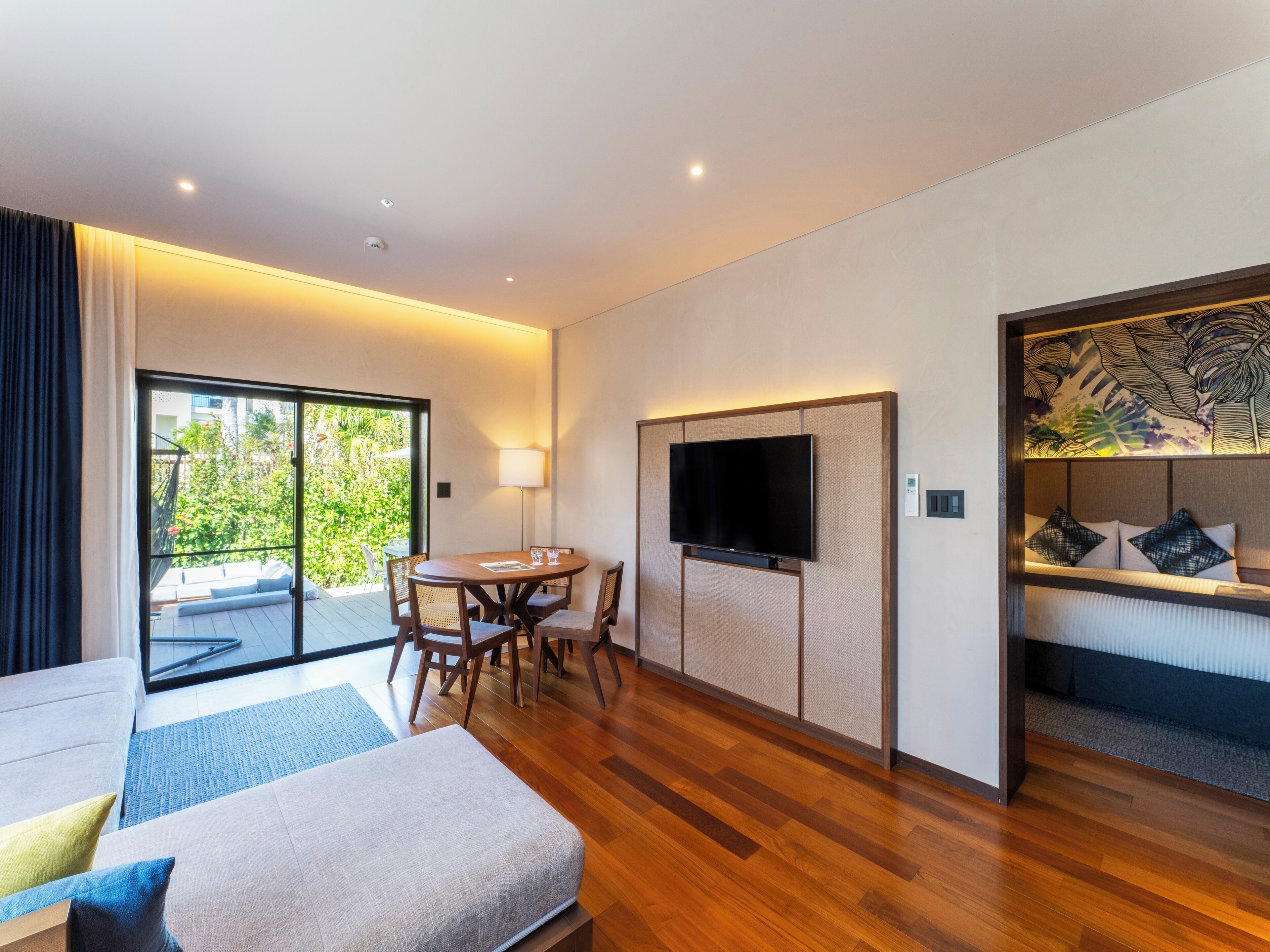 Een moderne hotelkamer met een houten vloer, een flatscreen televisie, een zithoek met een tafel en stoelen, en een balkon met uitzicht op groen.