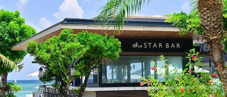 Un bar moderno con un letrero que dice “STAR BAR”, rodeado de exuberante follaje y con vista a un cuerpo de agua.