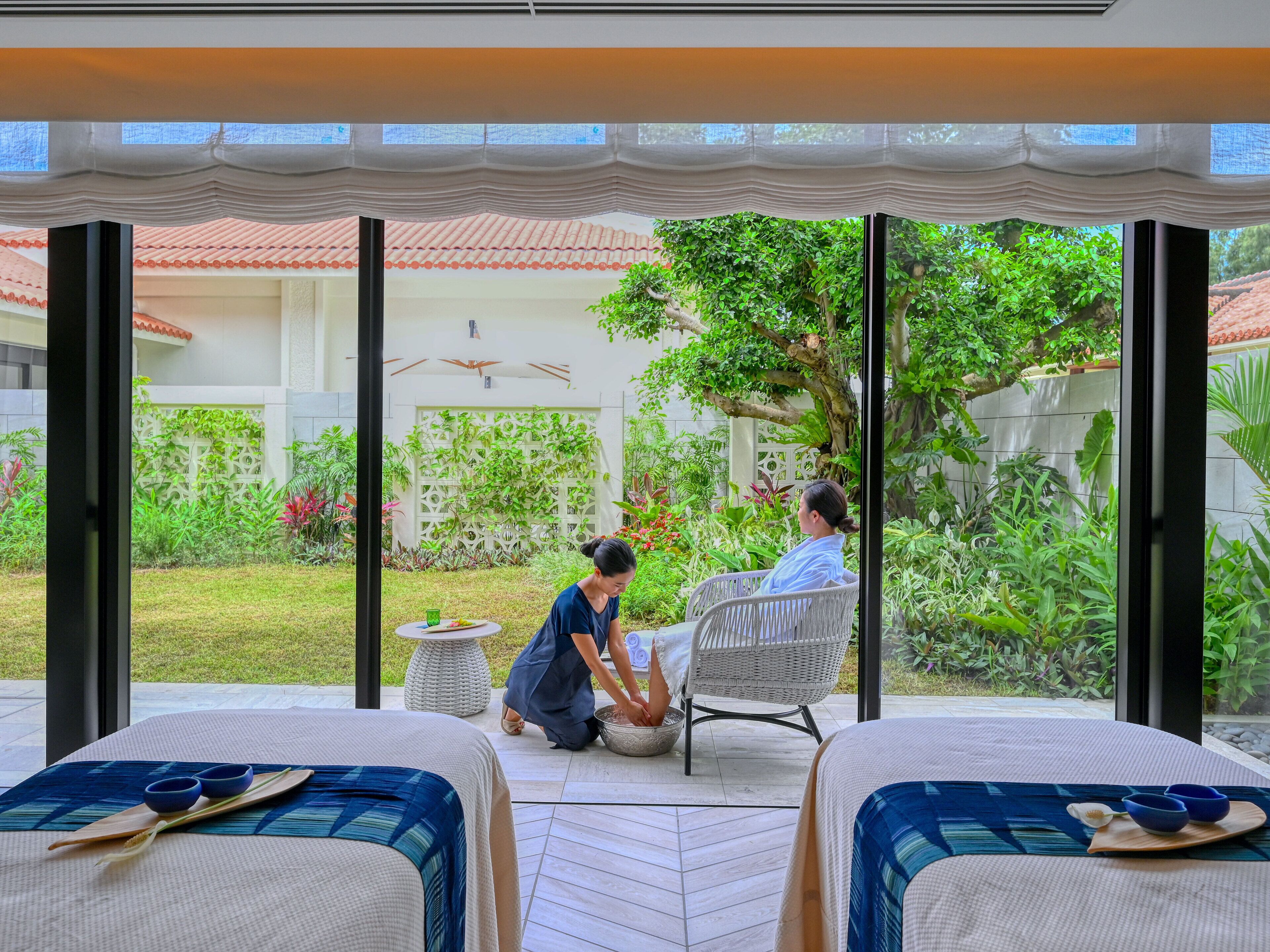 Una habitación con dos camas, una mesa de comedor y vistas a un jardín donde alguien cuida las plantas.